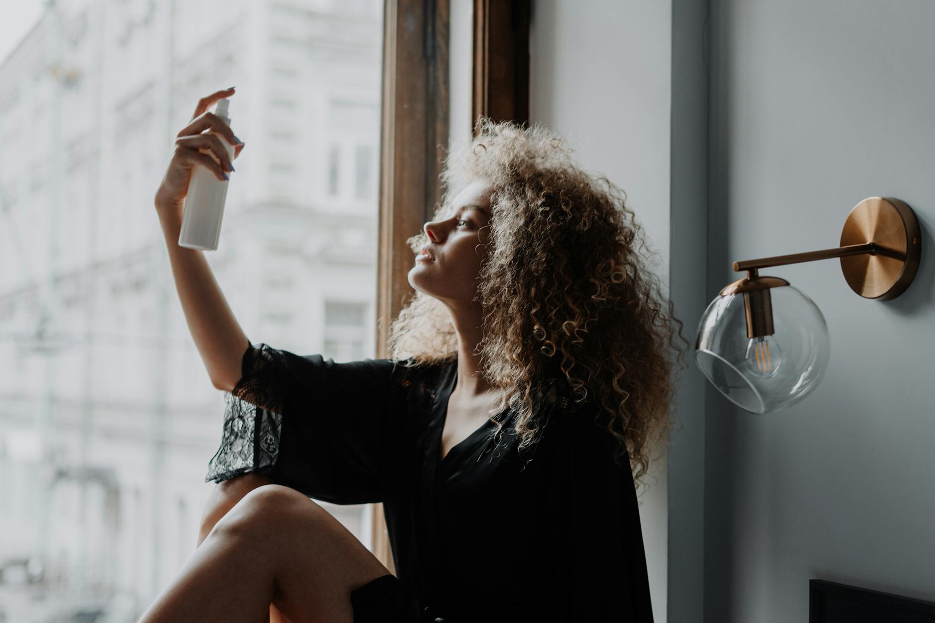 Kędzierzawa kobieta przy oknie psika włosy sprayem przeciw puszeniu