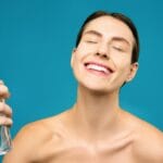 Uśmiechnięta kobieta spryskuje twarz mgiełką pielęgnacyjną na niebieskim tle