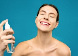 Uśmiechnięta kobieta spryskuje twarz mgiełką pielęgnacyjną na niebieskim tle